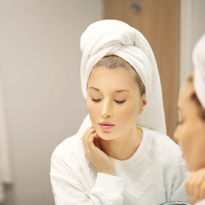 Žena s turbanem z ručníku se dívá do zrcadla a užívá si péči o pleť po aplikaci Krému Vitalní Rovnováhy.